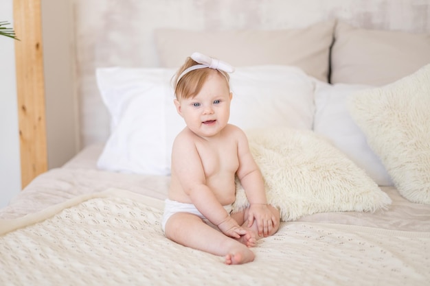 Uma menina encantadora em fraldas está sentada em um quarto brilhante em uma cama em casa Têxteis de quarto