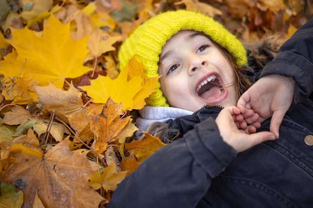 Uma menina engraçada com um chapéu amarelo em uma caminhada na floresta encontra-se na folhagem de outono.