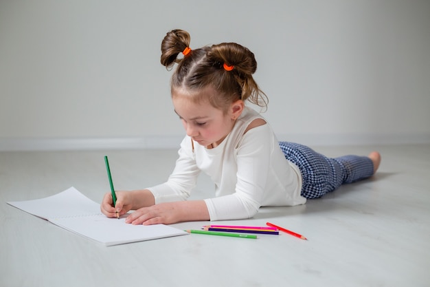 Uma menina engraçada está deitada no chão de madeira e desenhando em um álbum com lápis de cor