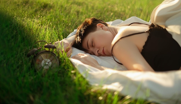 Uma menina envolta em um cobertor deita-se na grama verde ao lado de um despertador que promove um sono saudável