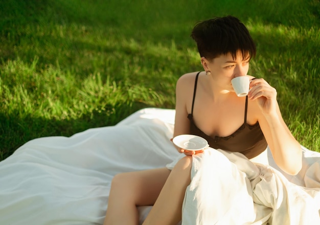 Uma menina envolta em um cobertor senta-se na grama verde com uma xícara de café desfrutando de uma manhã refrescante