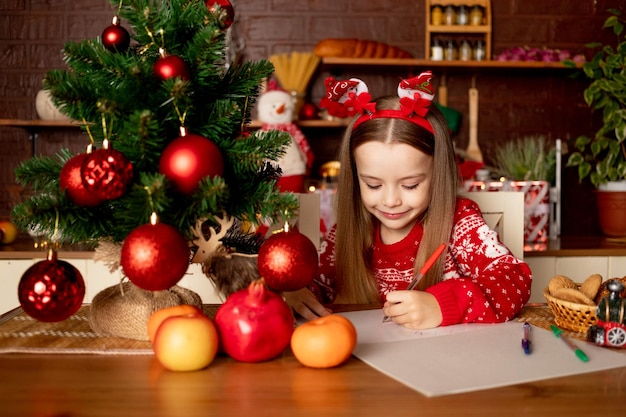 Uma menina escreve uma carta ao papai noel em uma cozinha escura debaixo de uma árvore de natal com bolas vermelhas o conceito de ano novo e natal