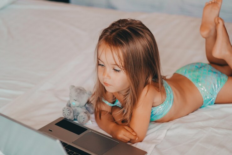 Uma menina está deitada em uma cama em um hotel com um ursinho de pelúcia cinza atrás de um laptop e assiste a desenhos animados