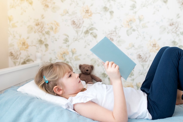 Uma menina está deitada na cama do quarto elegante e lê um livro azul, fazendo a lição de casa. Educação, conceito de ensino doméstico