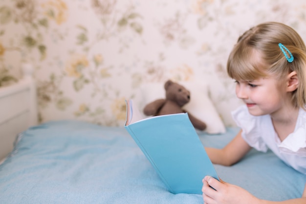 Uma menina está deitada na cama do quarto elegante e lê um livro azul, fazendo a lição de casa. Educação, conceito de ensino doméstico