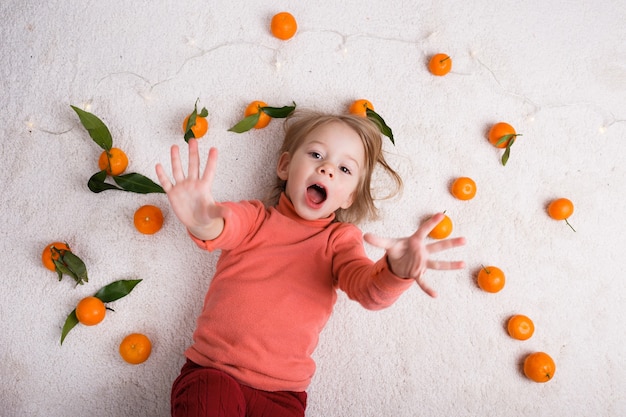 Uma menina está deitada no chão claro, um monte de tangerinas estão espalhadas ao seu redor.