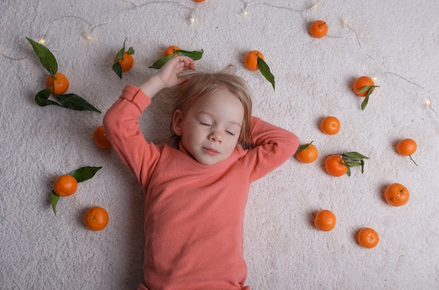 Uma menina está dormindo no chão claro, com um monte de tangerinas espalhadas ao seu redor.