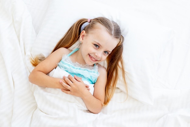 Uma menina está dormindo ou acordou de manhã em uma cama em casa em uma cama de algodão branco e está sorrindo docemente