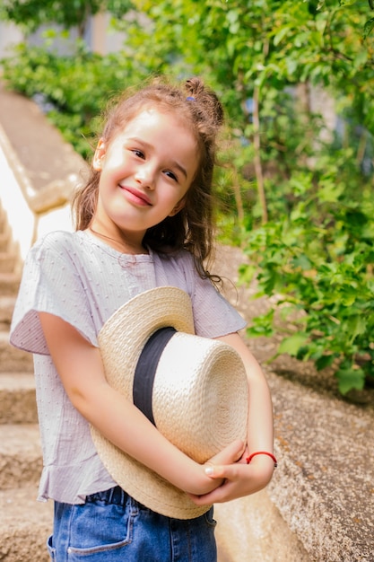 Uma menina está no parque e tem um chapéu nas mãos. A criança fica feliz e ri. Passeio de verão na natureza ou na cidade.