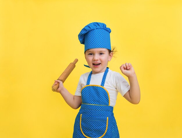 Uma menina está rindo em um avental de cozinha e chapéu com um rolo na mão em um espaço amarelo. Uma linda garota com um avental azul e amarelo e um chapéu de chef. Olhando .