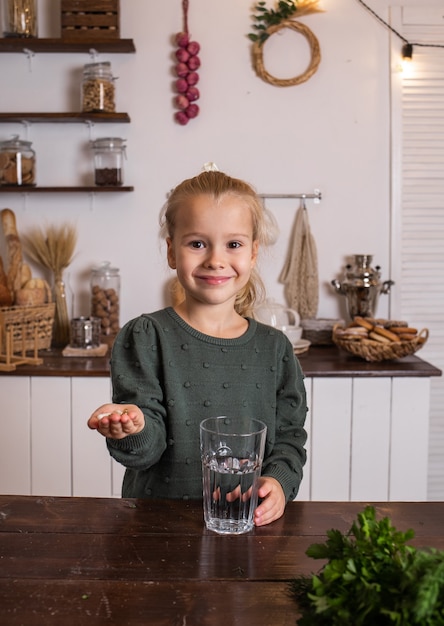 Uma menina está sentada à mesa da cozinha com um copo d'água e bebendo vitaminas