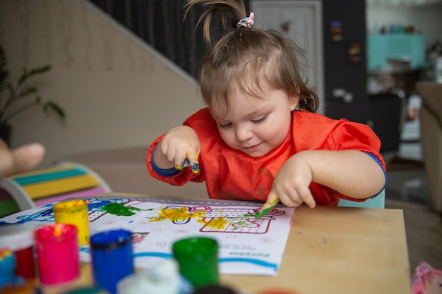 uma menina está sentada a uma mesa e desenhando com tintas a dedo. O conceito de desenvolvimento inicial