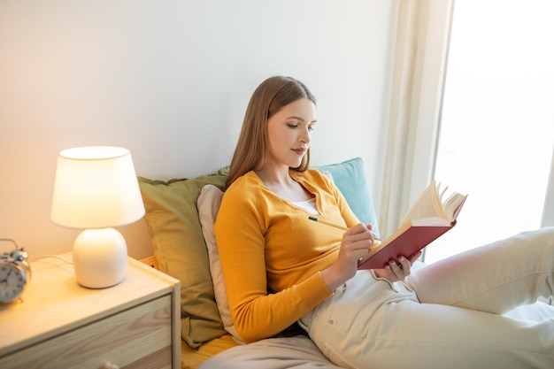 Uma menina está sentada em uma cama absorvida na leitura de um livro ela está vestida casualmente e parece focada em