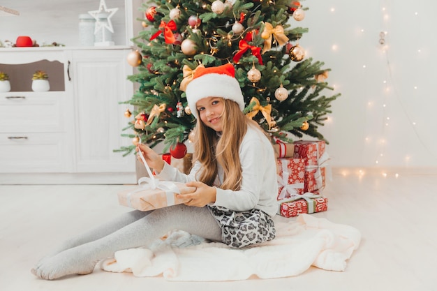 Uma menina está sentada perto da árvore de Natal com um presente de Natal. Feliz Natal.