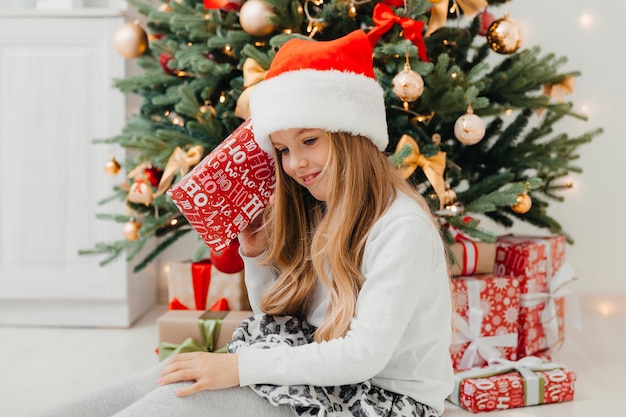 Uma menina está sentada perto da árvore de Natal com um presente de Natal. Feliz Natal.