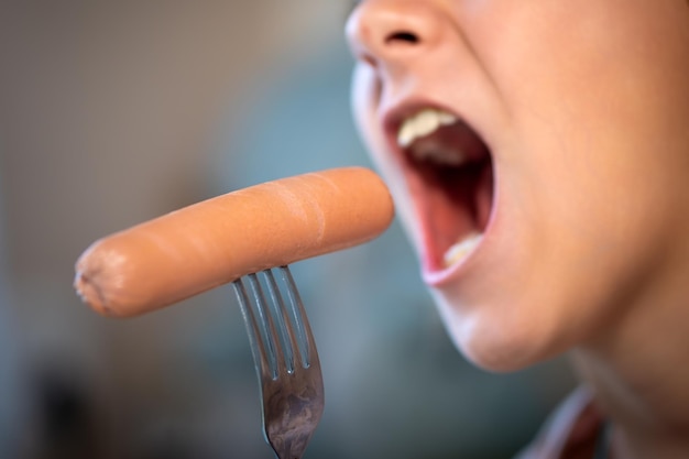 Uma menina faminta com a boca aberta come uma salsicha de um garfo deliciosa fast junk food Closeup