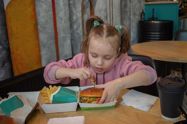 Foto uma menina faminta com apetite come fast-food em um café em uma mesa um hambúrguer e batatas fritas