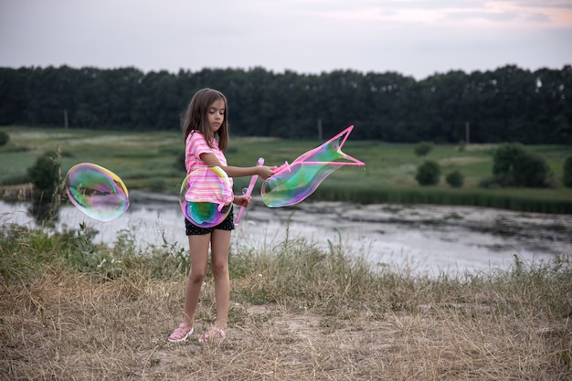 Uma menina faz grandes bolhas de sabão multicoloridas na natureza fora da cidade, descanso ativo.