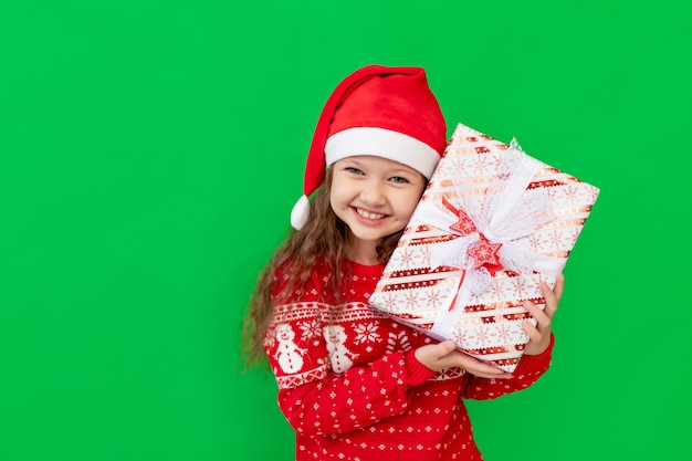 Uma menina feliz com roupas de inverno, um chapéu de Papai Noel e um suéter sobre um fundo verde isolado oferece presentes para o ano novo ou Natal e sorri, um lugar ou espaço para texto