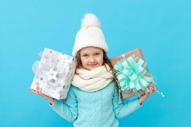 Uma menina feliz com roupas de inverno, um chapéu e um suéter sobre um fundo azul isolado oferece presentes para o ano novo ou natal e sorri, um lugar ou espaço para texto