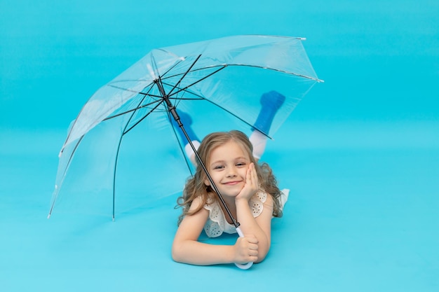 Uma menina feliz e fofa com botas de borracha azuis e um vestido de algodão branco segurando um guarda-chuva deitada sobre um fundo azul no estúdio rindo, sorrindo e brincando, um lugar para texto