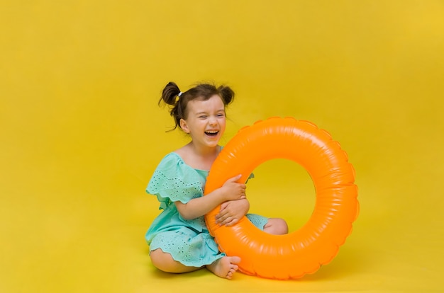 Uma menina feliz em um vestido azul se senta com um círculo inflável e ri em um fundo amarelo com uma cópia do espaço. Férias de verão
