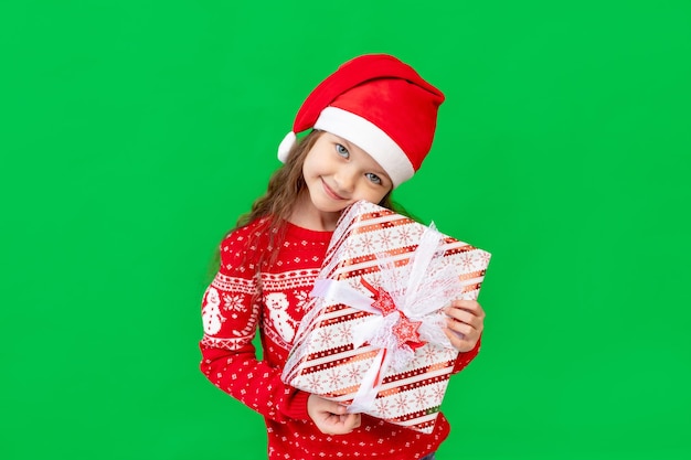 Uma menina feliz no inverno veste um chapéu de Papai Noel e um suéter em um fundo verde isolado mantém presentes para o ano novo ou Natal e sorri um lugar ou espaço para texto