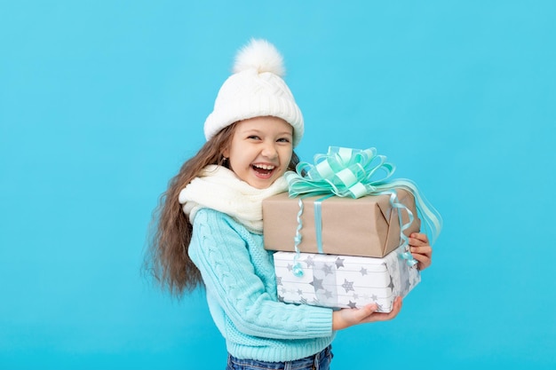 Uma menina feliz no inverno veste um chapéu e um suéter em um fundo azul isolado mantém presentes para o ano novo ou Natal e sorri um lugar ou espaço para texto