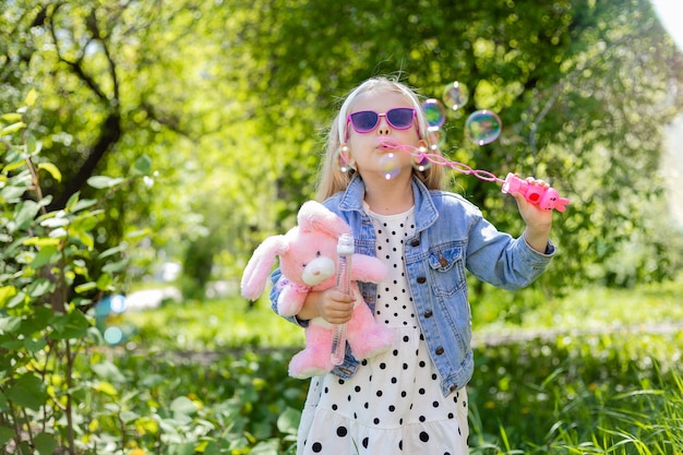 Uma menina feliz no verão com óculos de sol inflando bolhas de sabão e segurando um brinquedo nas mãos