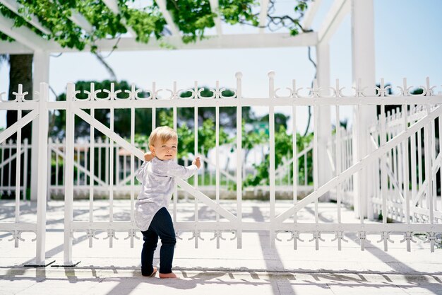 Uma menina fica perto de uma cerca de metal branco entrelaçada com vegetação em um parque