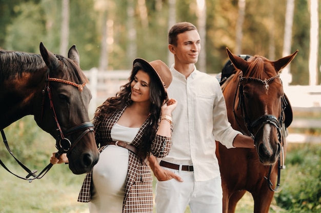 Uma menina grávida com um chapéu e o marido com roupas brancas estão ao lado de cavalos na floresta na natureza. Mulher grávida elegante com um homem com cavalos. Família.