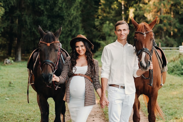 Uma menina grávida com um chapéu e o marido com roupas brancas estão ao lado de cavalos na floresta na natureza. Mulher grávida elegante com um homem com cavalos. Família.