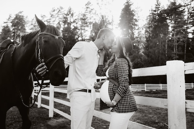 Uma menina grávida com um chapéu e o marido com roupas brancas ficam ao lado dos cavalos perto do curral de cavalos ao pôr do sol. Mulher grávida elegante com um homem com cavalos. foto em preto e branco.