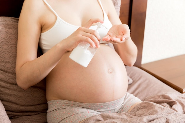 Uma menina grávida senta-se em casa na cama e esfrega sorriso um creme anti-estrias no estômago Preparação da maternidade da gravidez e conceito de expectativa