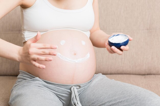 Foto uma menina grávida senta-se em casa na cama e esfrega sorrisos um creme anti-marcas de estiramento em seu estômago gravidez preparação para a maternidade e conceito de expectativa