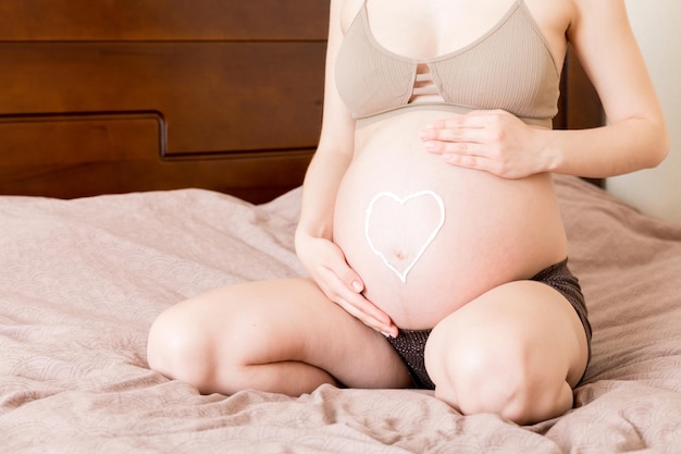 uma menina grávida senta-se em casa na cama e passa no coração um creme anti-estrias na barriga Gravidez, preparação para a maternidade e conceito de expectativa