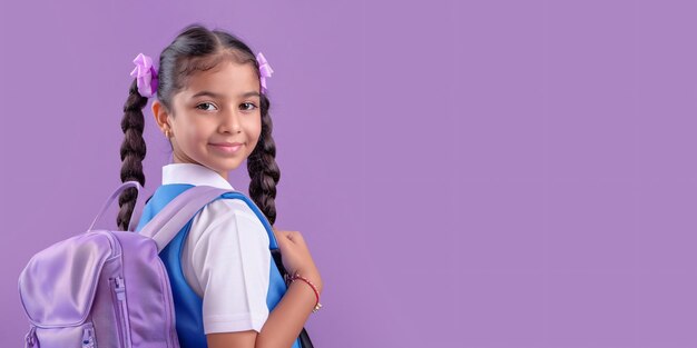 Uma menina indiana pré-adolescente alegre com uma criança hispânica em trajes escolares segurando uma mochila de pé sozinha contra um fundo roxo olhando para o espectador representando o início de um novo ano escolar