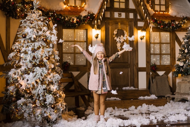 Uma menina levanta as mãos e borrifa neve no fundo de uma casa de Natal e uma árvore de Natal coberta de neve artificial