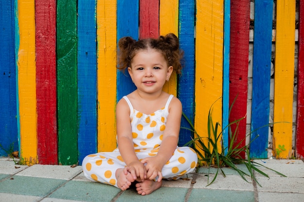 Uma menina linda sentada perto de uma cerca de madeira multicolorida