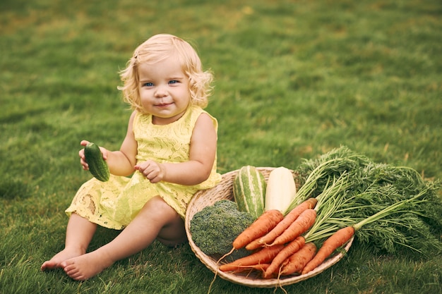 Uma menina loira em um vestido amarelo, com os pés descalços, tem um pepino na mão, senta-se ao lado de uma grande cesta de legumes na grama. comida saudável, comida vegetariana verde.