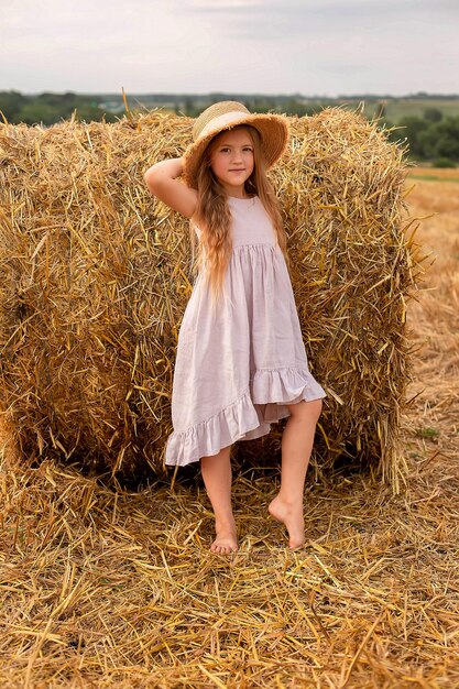 uma menina loira em um vestido de linho rosa e um chapéu de palha fica ao lado de um palheiro em um campo ceifado