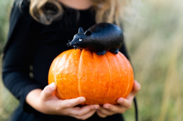 Foto uma menina loira em uma fantasia preta e um chapéu de bruxa, halloween com uma abóbora e um rato nas mãos, uma criança alegre.