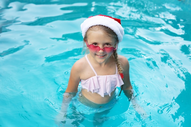 uma menina loira está de pé na piscina com um chapéu de Papai Noel e óculos cor de rosa sorrindo no ano novo