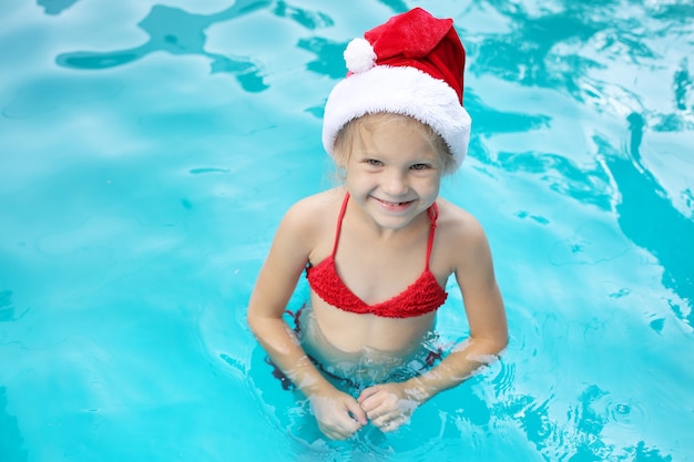 uma menina loira está de pé na piscina com um chapéu de Papai Noel e óculos cor de rosa sorrindo no ano novo