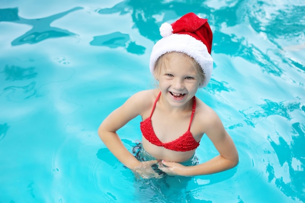 uma menina loira está de pé na piscina com um chapéu de Papai Noel e óculos cor de rosa sorrindo no ano novo