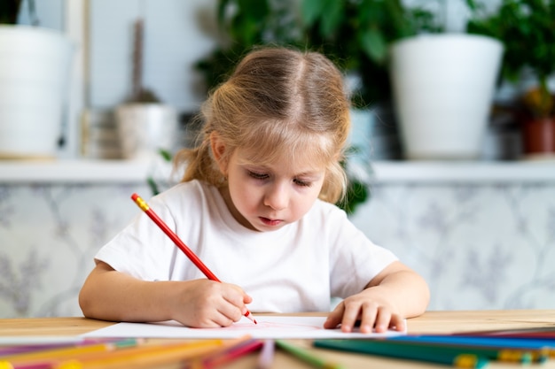 uma menina loira está sentada à mesa, sorrindo e desenhando com um lápis vermelho, ali