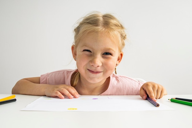 Uma menina loira sorridente desenha com lápis de cor sentado em uma mesa branca