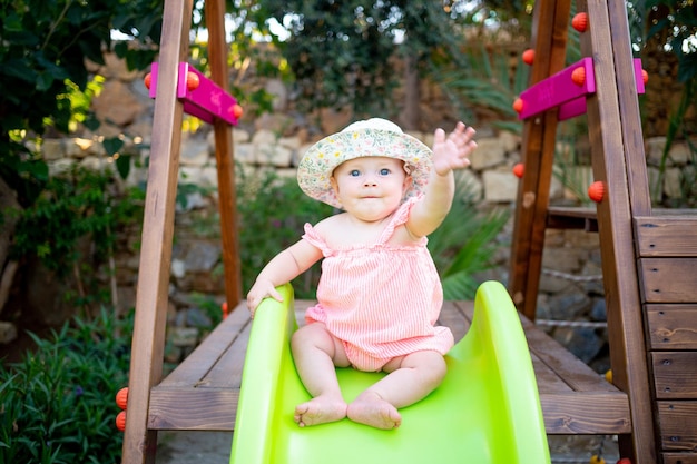 Uma menina monta um slide no playground e ri no verão com um chapéu panamá e pés descalços