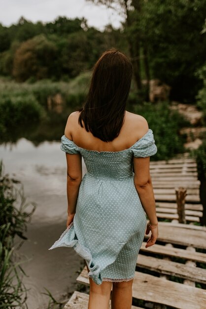 Uma menina morena bronzeada com um vestido turquesa atravessa sozinha o rio em uma velha ponte de madeira.