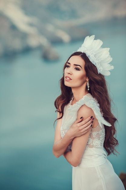 Uma menina morena com um lindo vestido branco, como um anjo em um penhasco acima do mar, dança ao pôr do sol.